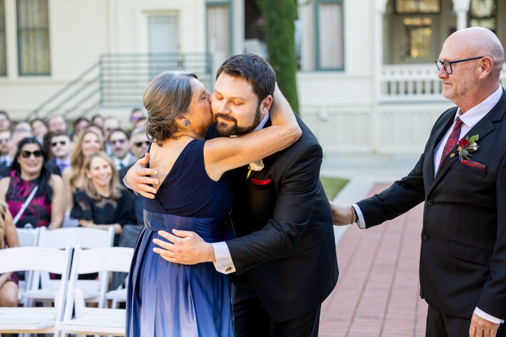 Two people share an emotional embrace at an outdoor wedding ceremony while guests look on from white chairs.