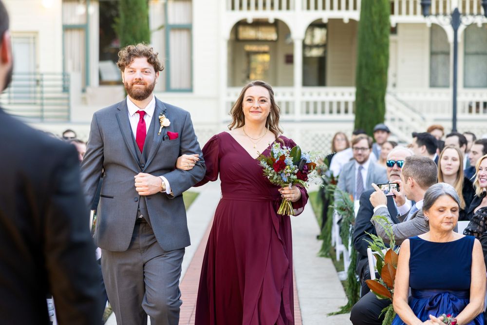 A wedding party stands in formal attire with burgundy and navy dresses and gray suits at an outdoor venue.