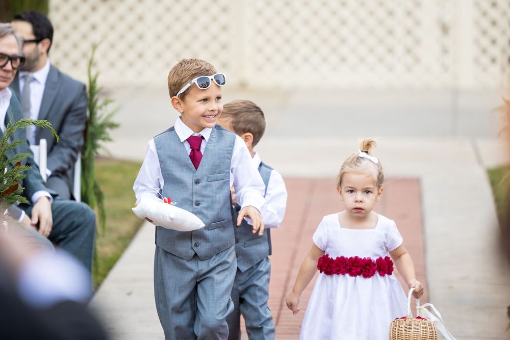 Young ring bearer in gray vest and red tie walks down aisle with flower girl in white and red dress at outdoor wedding.