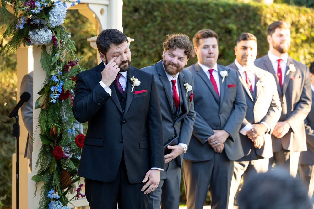 Wedding party in black suits and red ties standing together in front of a decorated outdoor gazebo.