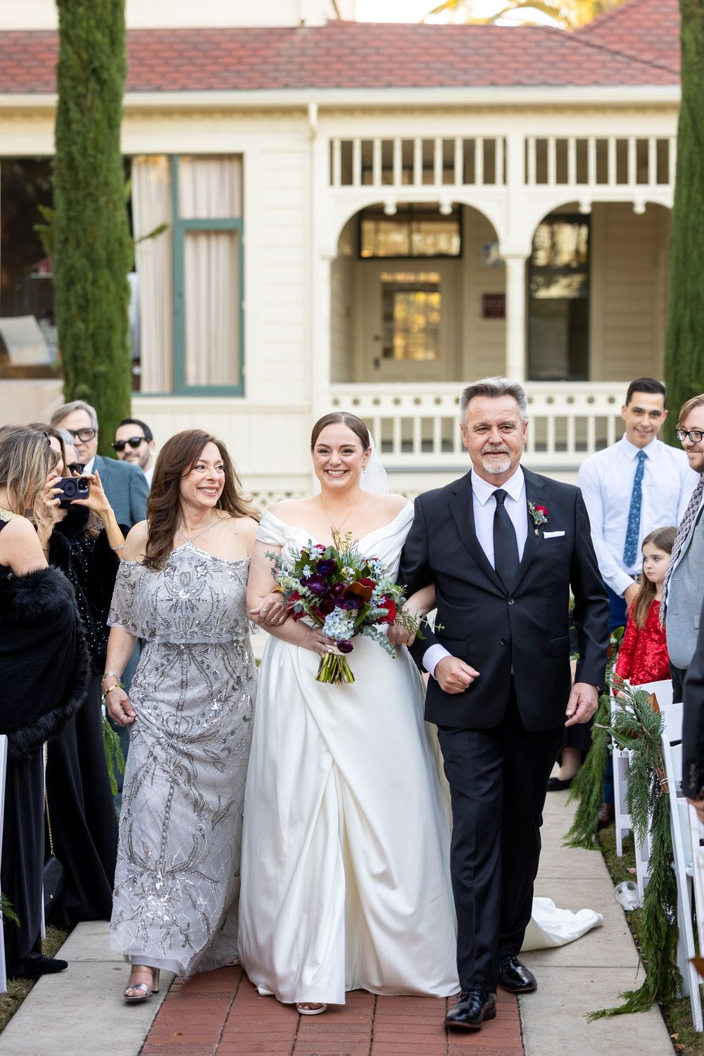 Wedding celebration on a brick walkway with formal attire and guests taking photos along the sides.