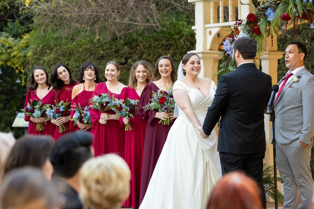 Wedding party members in burgundy dresses and gray suits gather for outdoor ceremony photos at elegant venue.