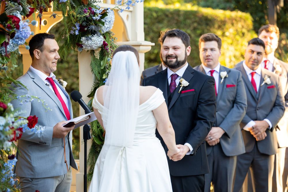 Wedding ceremony taking place outdoors with wedding party in grey suits and red ties standing at the altar.