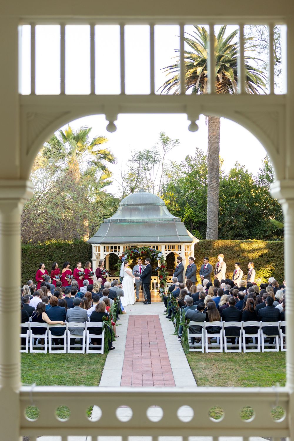 Outdoor wedding ceremony under a gazebo viewed through an ornate white archway with palm trees in background.