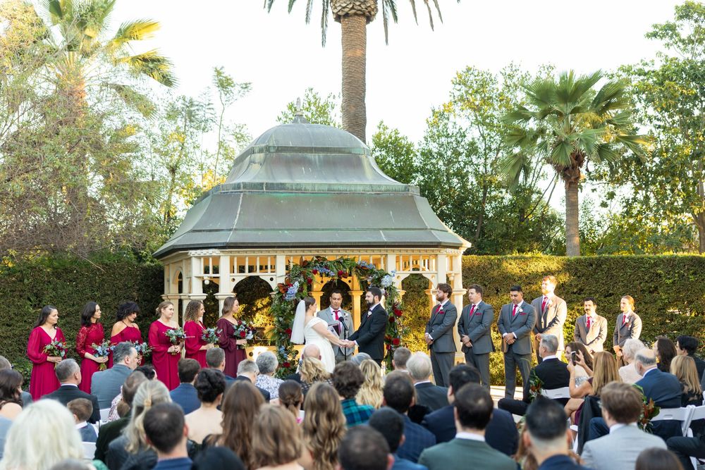 Large outdoor wedding ceremony with guests seated around a central gazebo surrounded by palm trees.