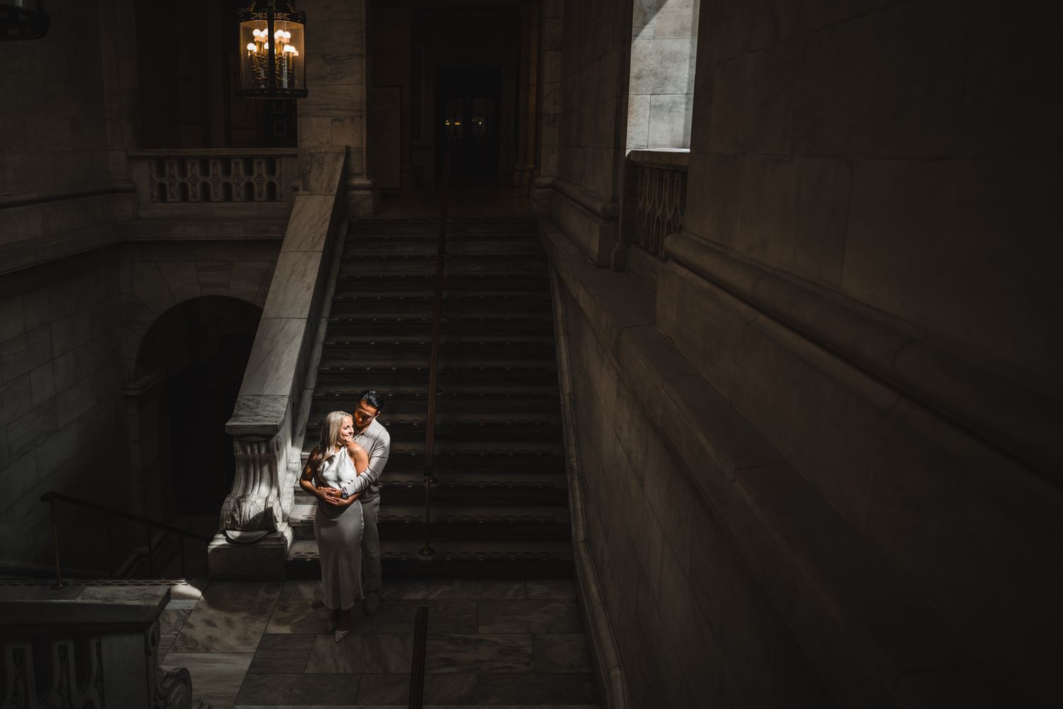 A couple shares an intimate moment in a dimly lit stairwell with dramatic shadows and lighting.