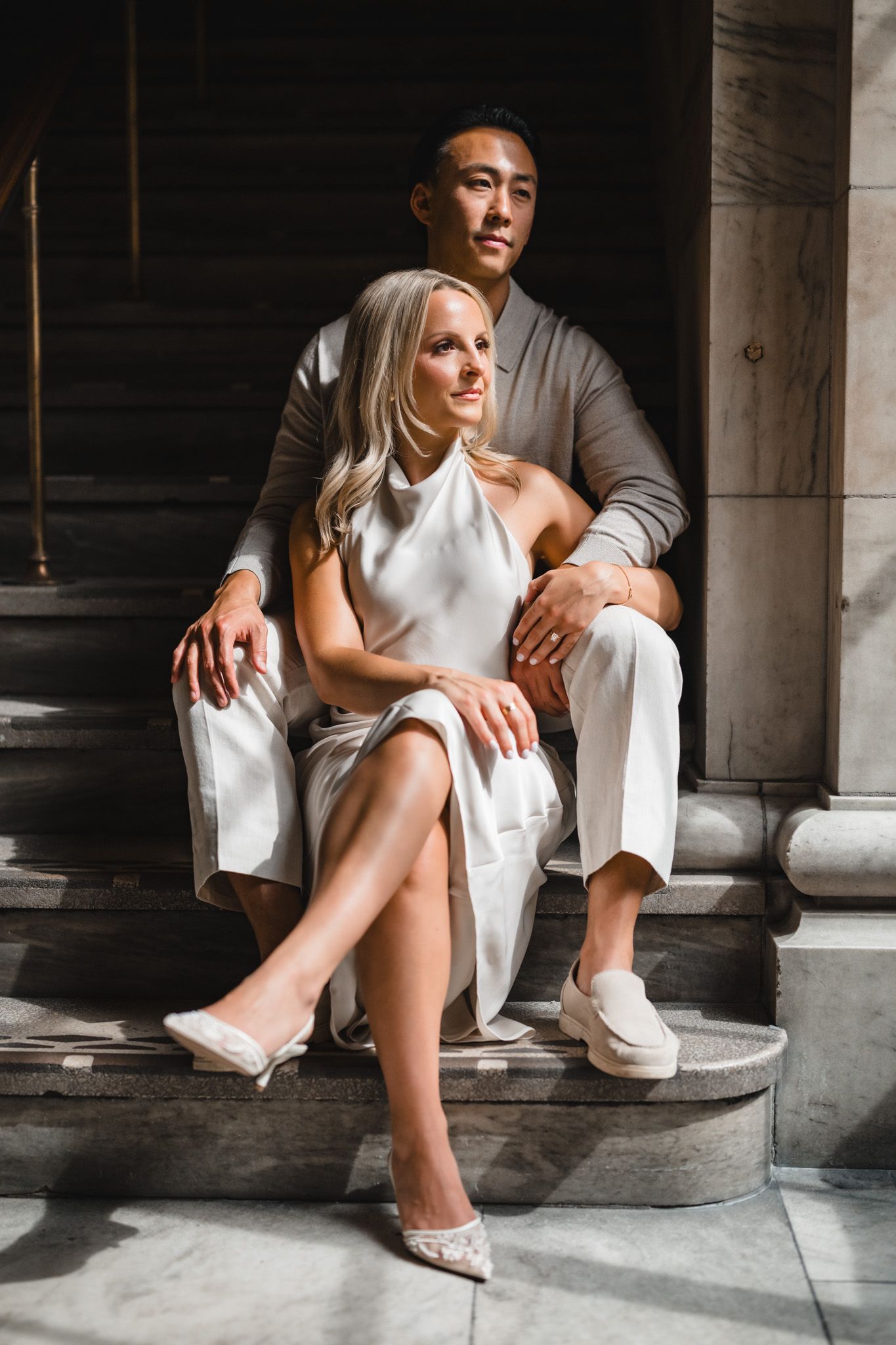 A couple in white outfits sits together on stone steps in an architectural setting.
