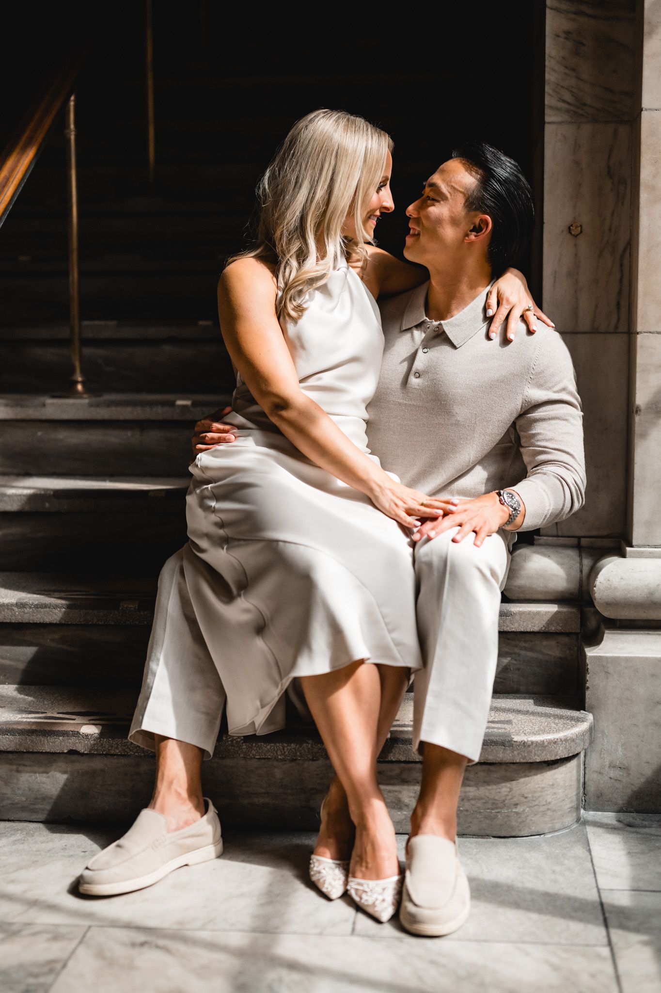 Two people in light colored clothing sit close together on stone steps with dramatic lighting.