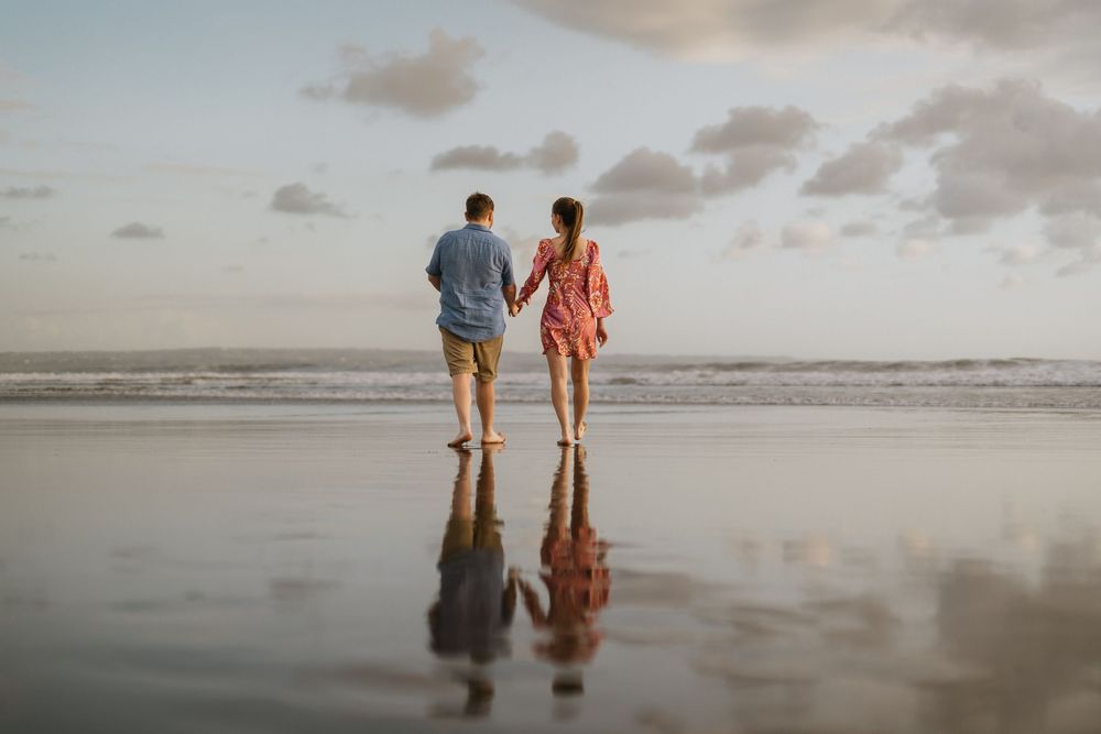 Two people walking hand in hand on a reflective beach at sunset with their silhouettes mirrored in the wet sand.