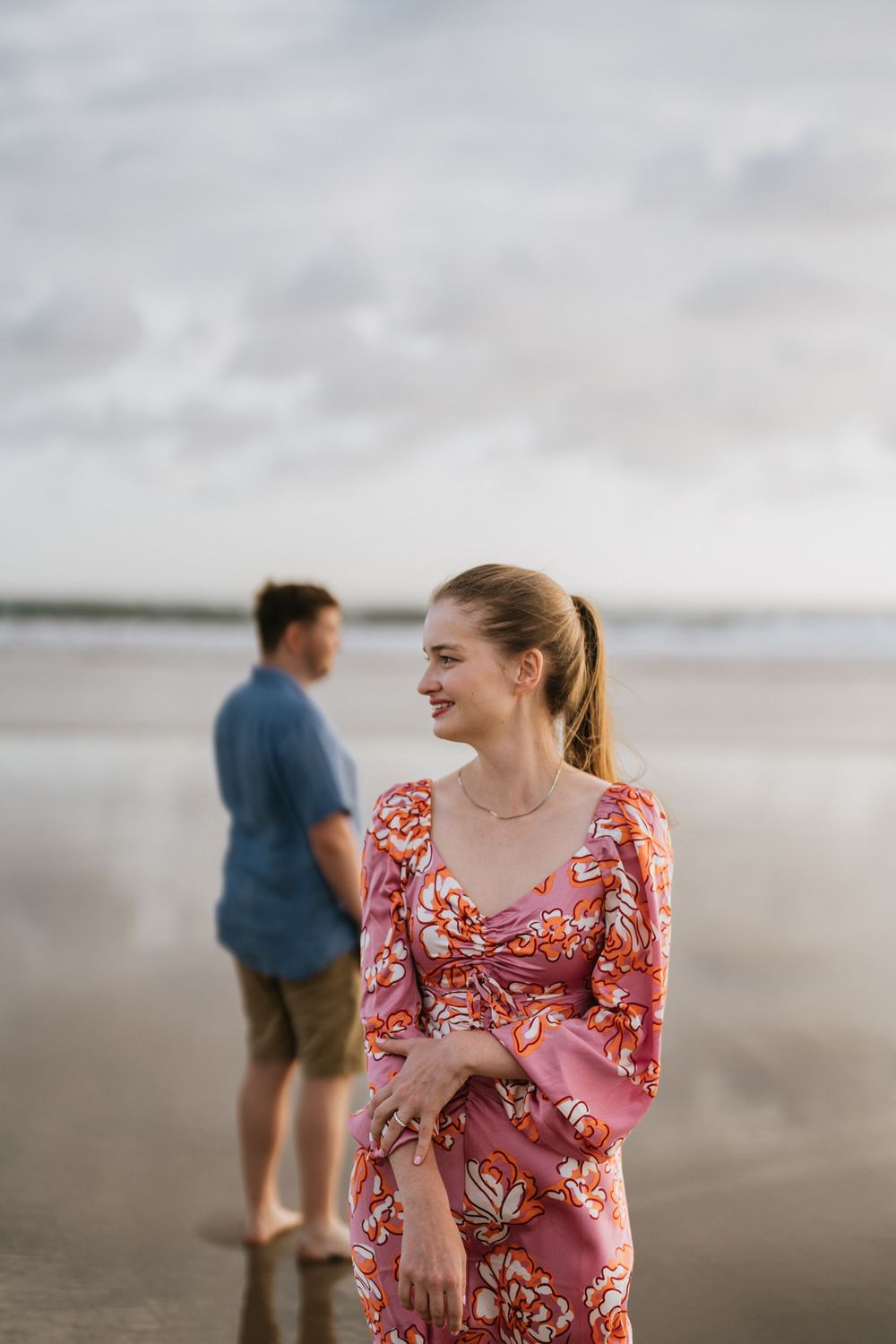A couple stands on a cloudy beach, with a woman in a pink floral dress smiling while someone stands in the background.