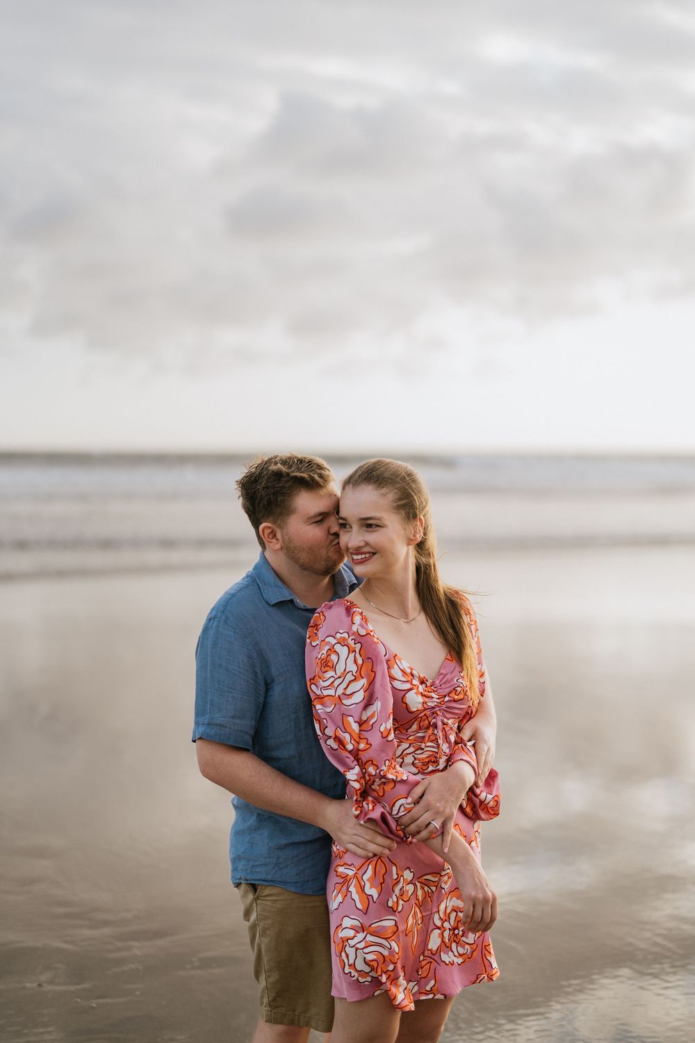 A romantic couple embraces on a misty beach, with her wearing a pink floral dress and him in a blue shirt.