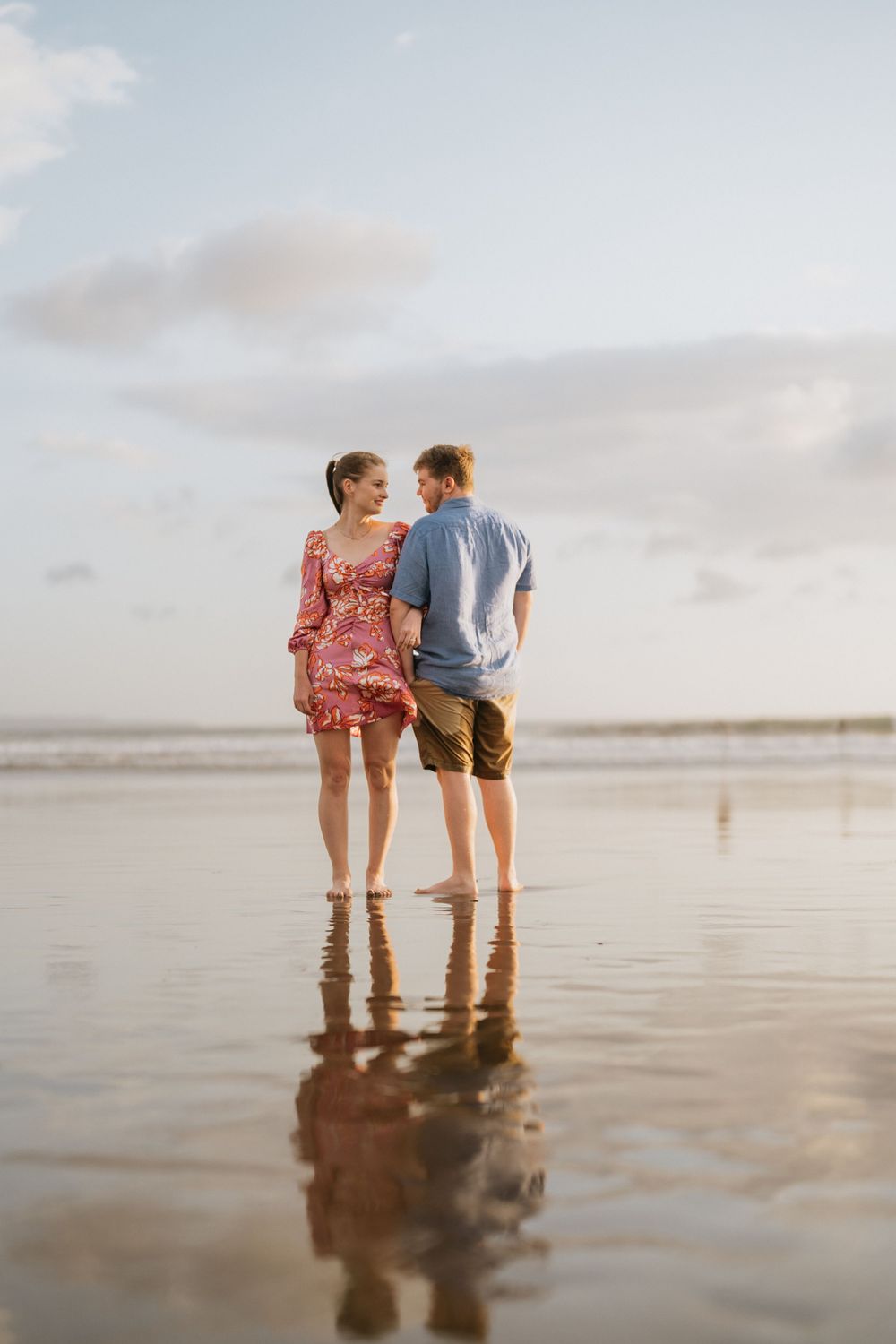 A couple stands together on a reflective beach at sunset creating mirror-like reflections in the wet sand.