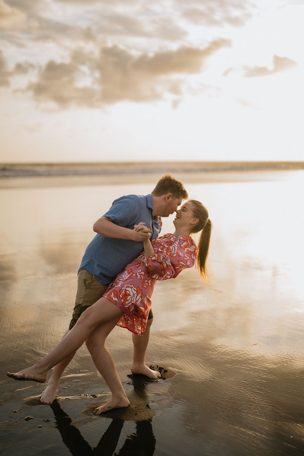 A couple shares a romantic moment while dipping on a beach at sunset, creating a silhouette against the golden sky.