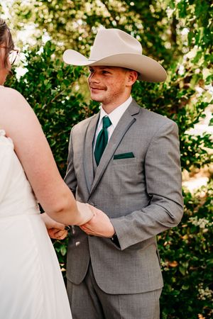 A groom in a cowboy hat and grey suit smiles during an outdoor wedding ceremony.