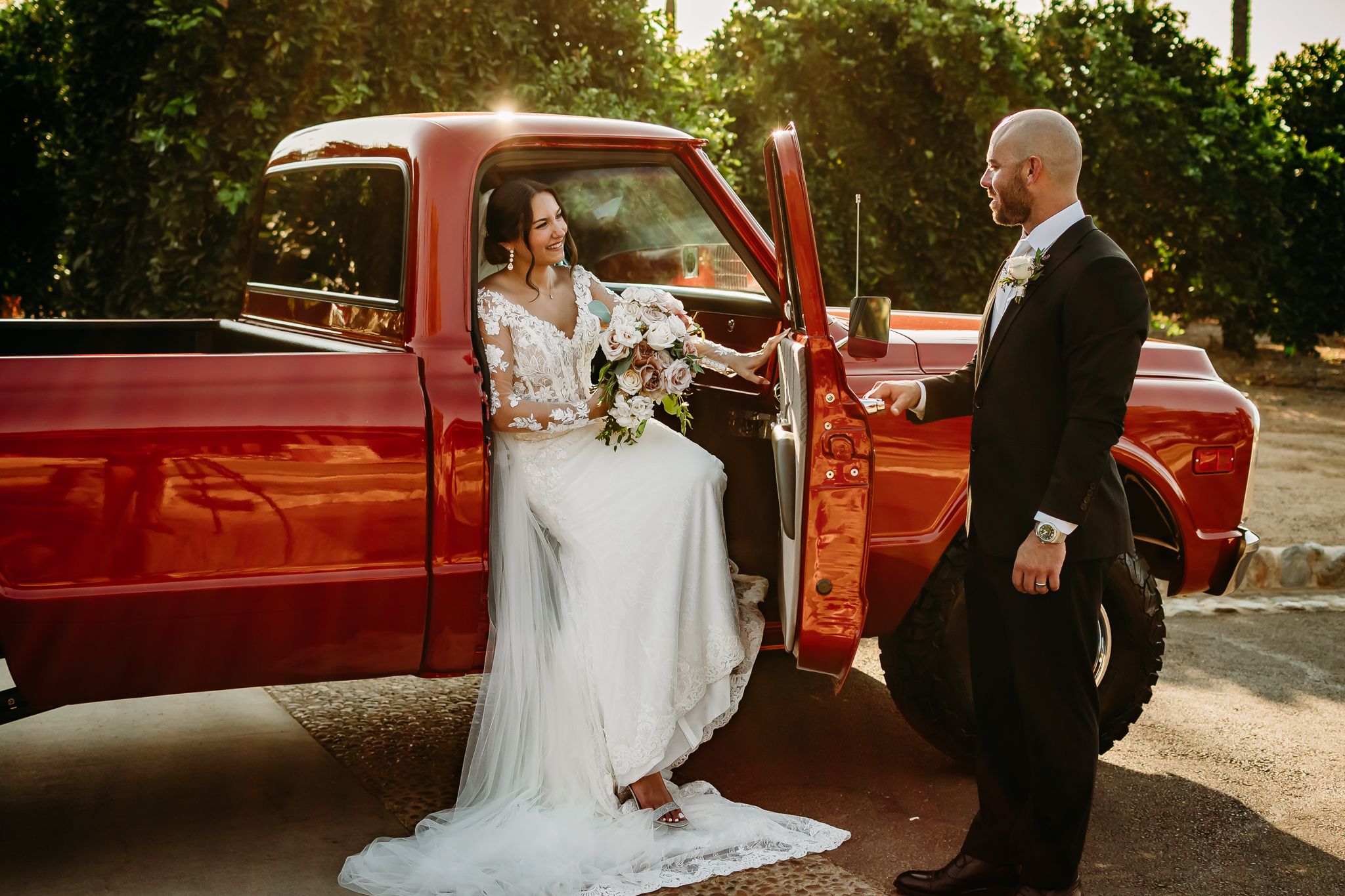 A bride in white dress sits in a vintage red pickup truck on her wedding day.