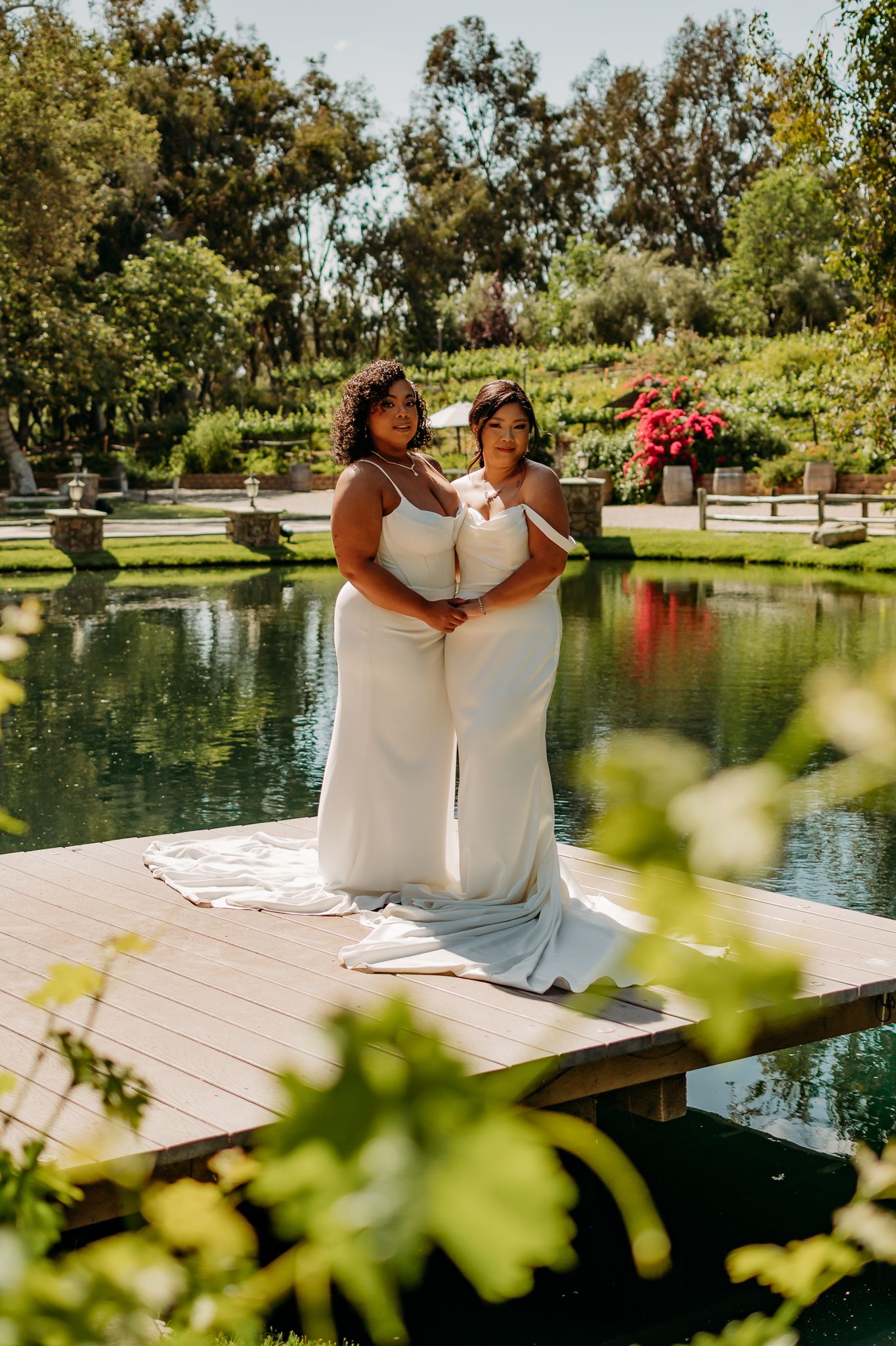 Two brides in white dresses share a tender moment on a dock by a garden pond.
