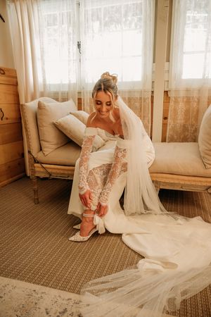 A bride in a lace wedding dress adjusts her shoe in a sunlit room.