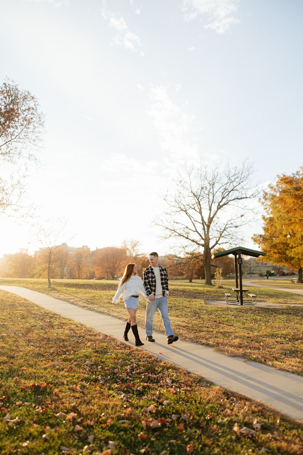 Couple walks along autumn path with fallen leaves and golden sunlight.