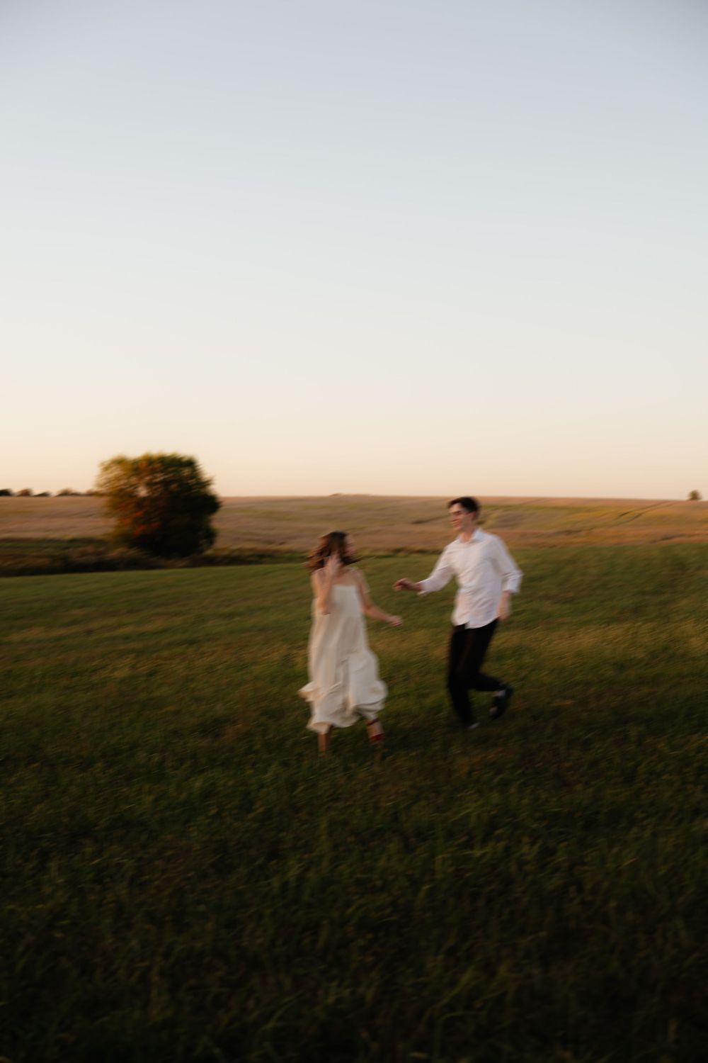 Couple running playfully through grassy field at sunset wearing white dress and shirt.