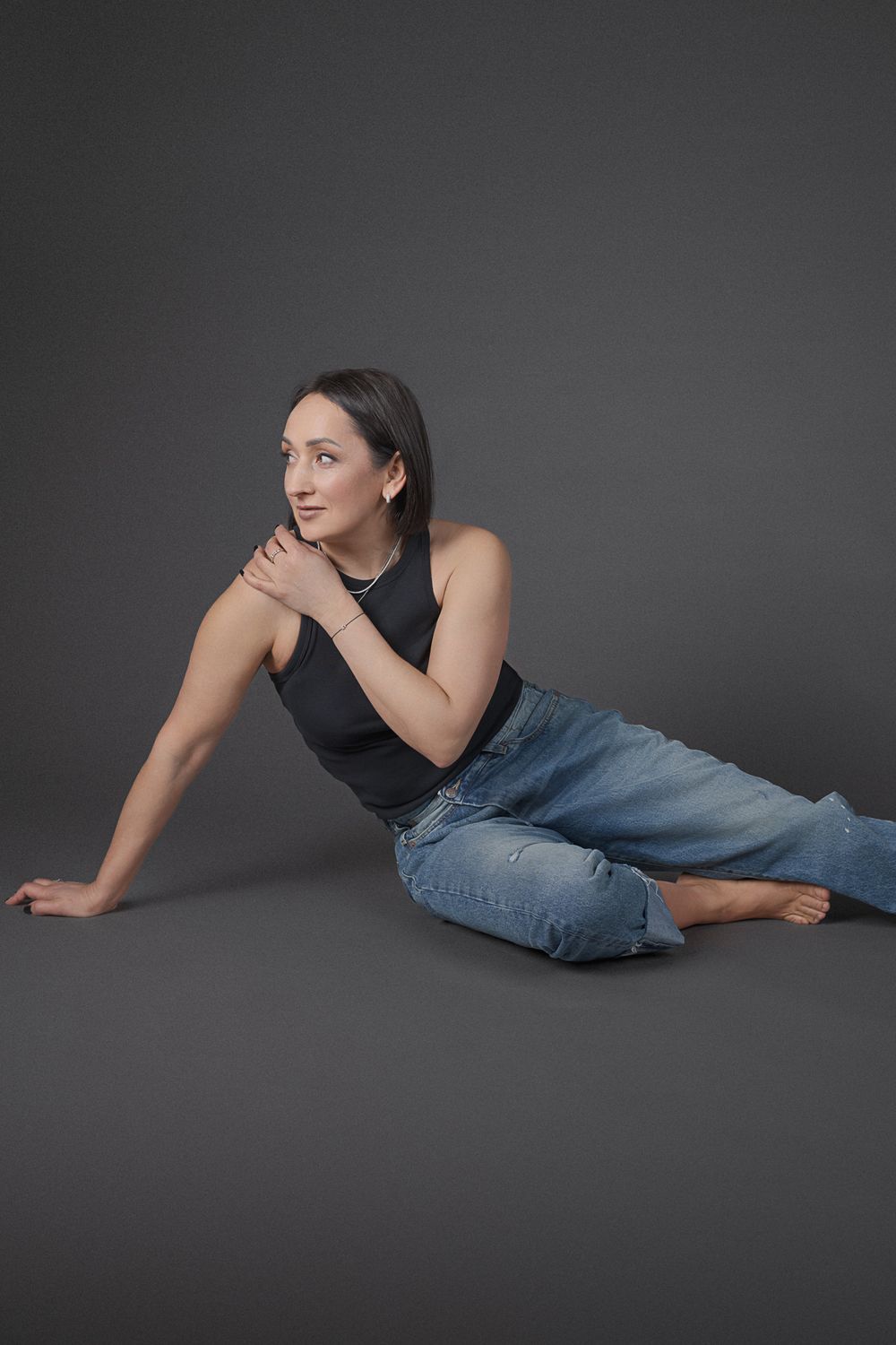 Artistic fashion poses on floor wearing black top and blue jeans against dark background.
