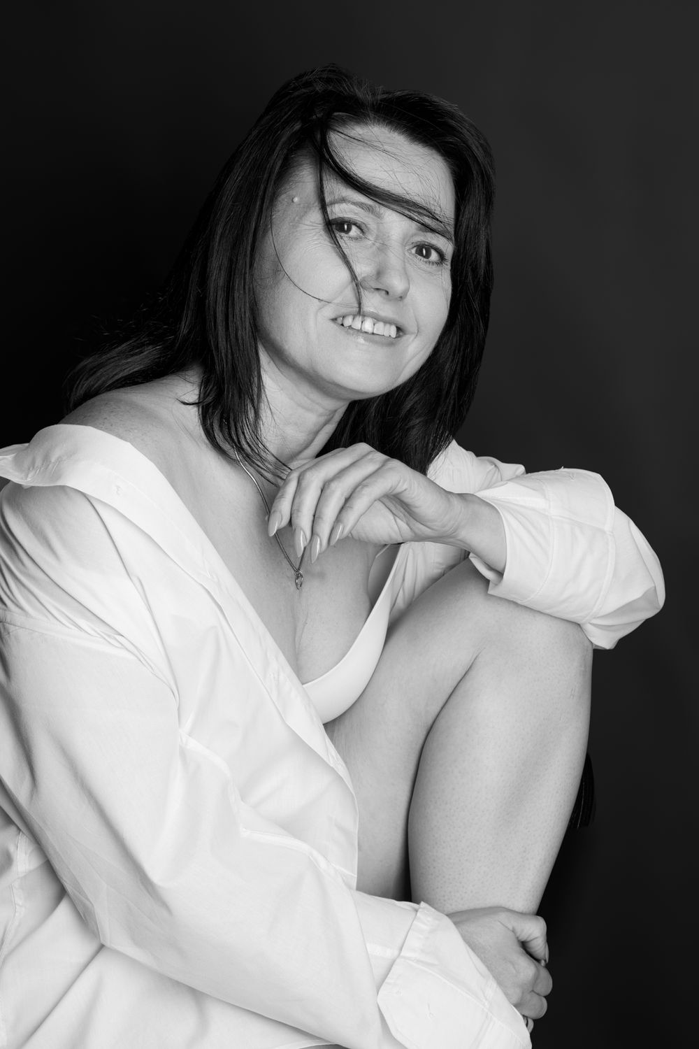 Black and white portrait of a dark-haired woman in a white shirt, sitting with knees up, smiling against dark background.