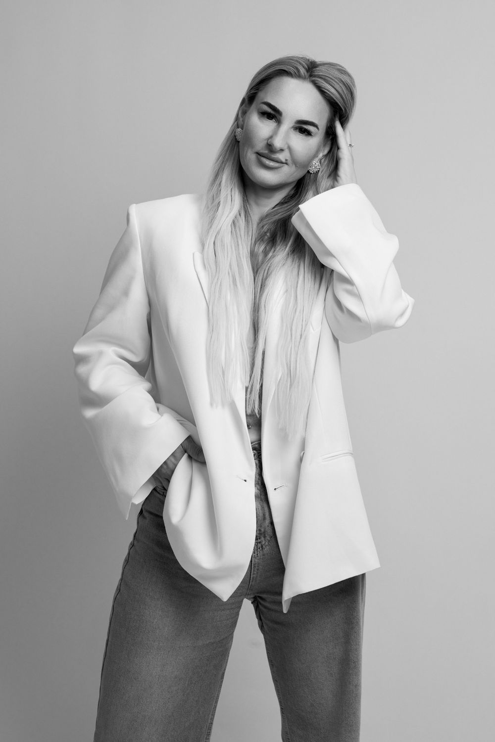 Black and white portrait of a blonde woman in a white blazer and jeans posing confidently against a gray background.