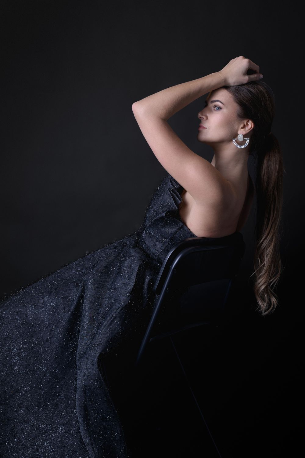 Woman in elegant black strapless gown with crystal earrings poses gracefully against dark background.