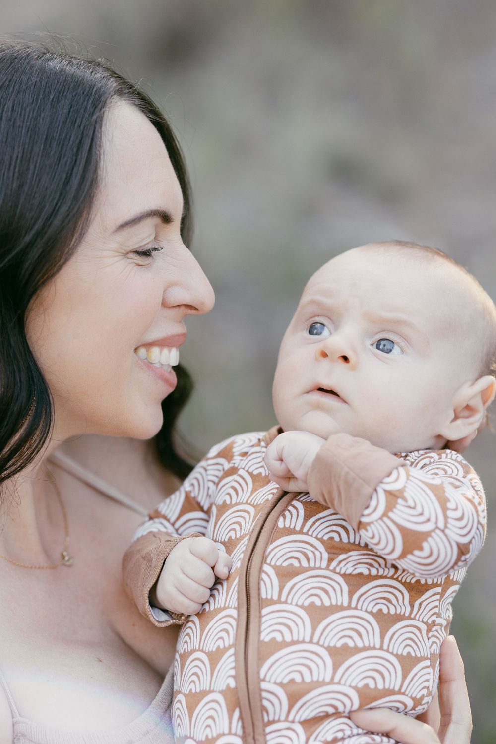 Smiling mother holding alert blue-eyed baby wearing brown rainbow print zip-up outdoors.