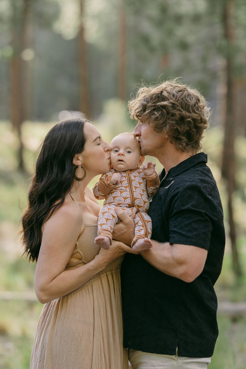 Loving parents kissing their newborn baby together in a sunlit forest setting.