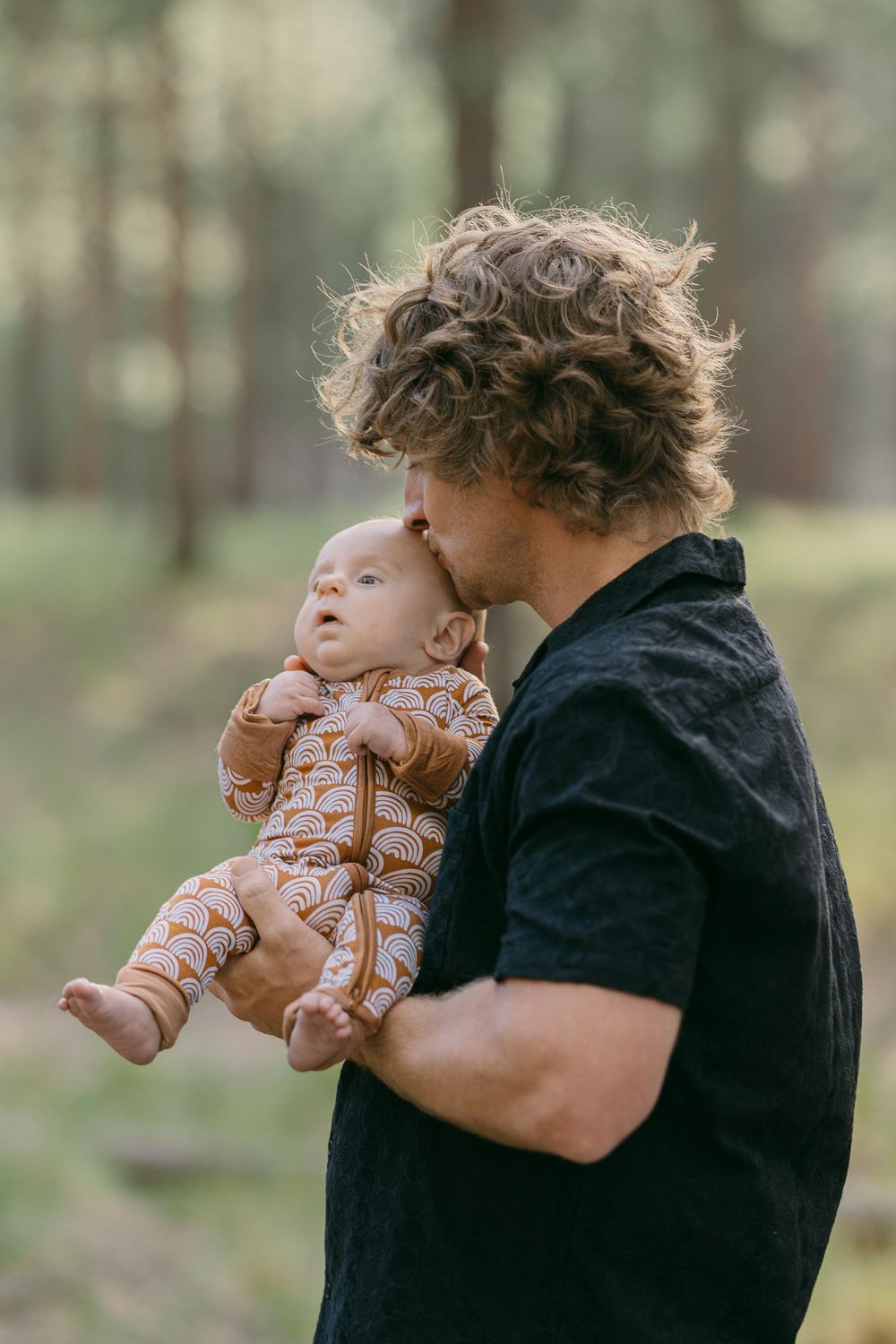 Curly-haired father tenderly kissing his newborn baby outdoors in a pine forest.