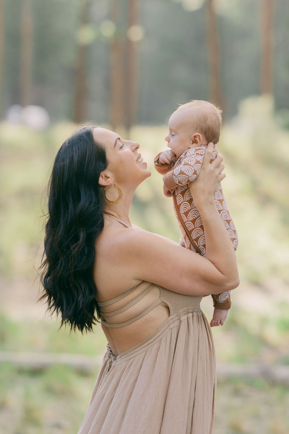 Joyful dark-haired mother in beige dress lifting smiling newborn baby in sunlit forest.