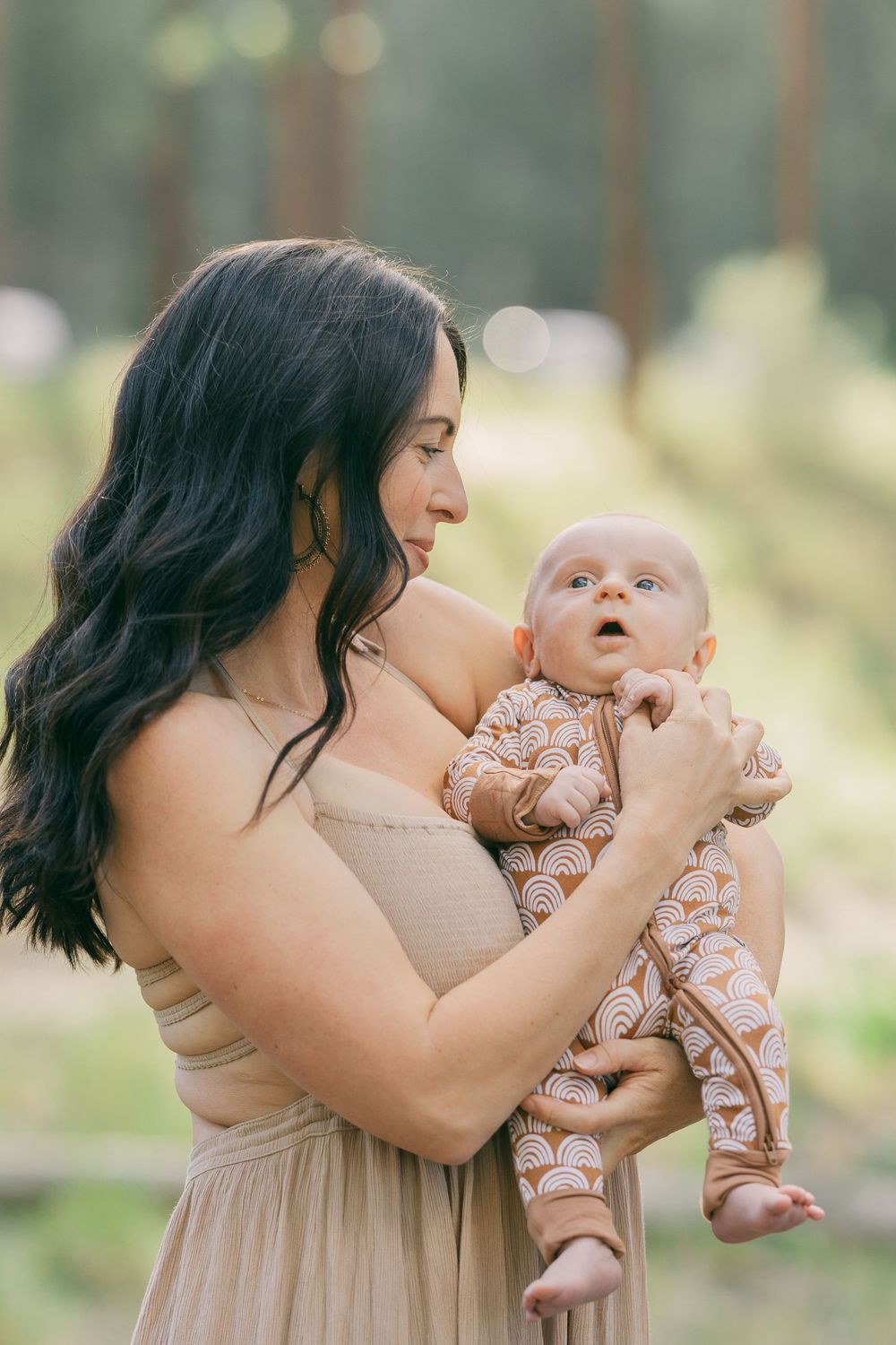 Dark-haired mother in elegant beige dress holding wide-eyed newborn baby in forest setting.