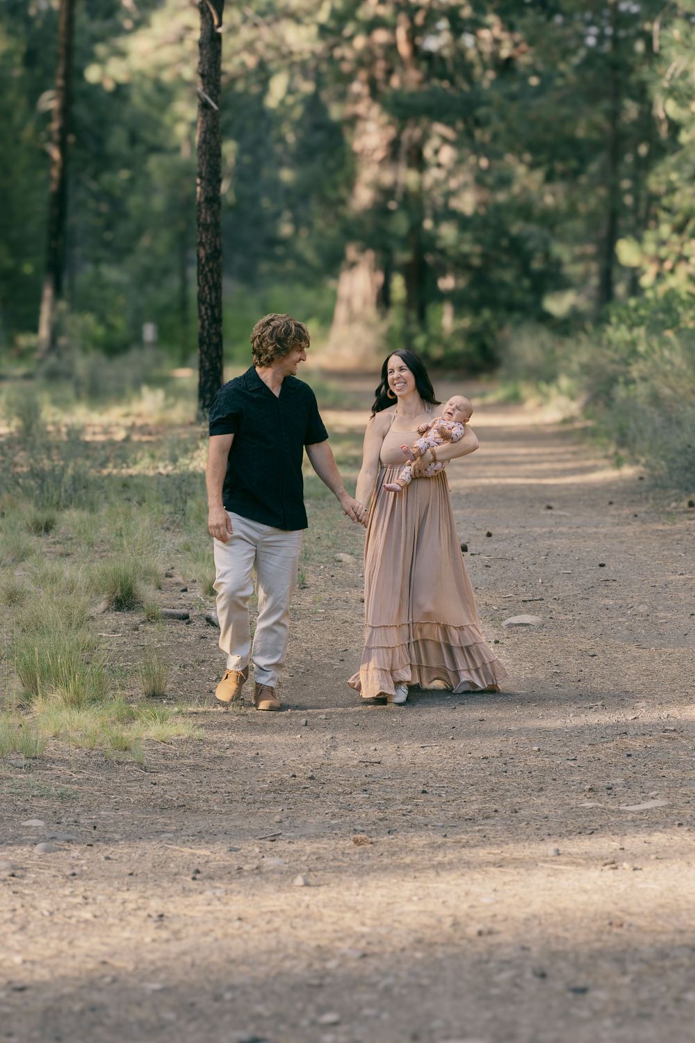 Young family strolling hand-in-hand on forest path, mother carrying newborn baby in patterned outfit.