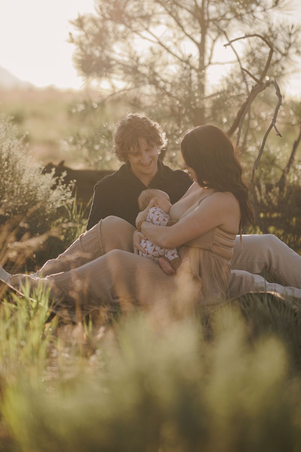 Young parents sitting in sun-drenched meadow together, tenderly cradling their newborn baby.