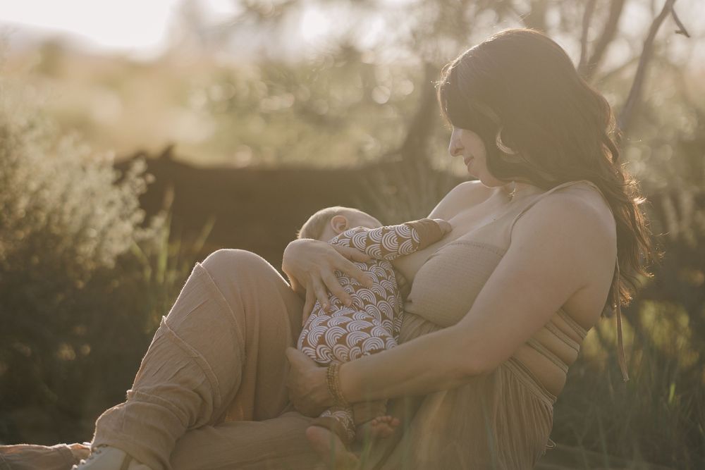 Mother breastfeeding newborn baby outdoors in golden sunlight among green foliage.