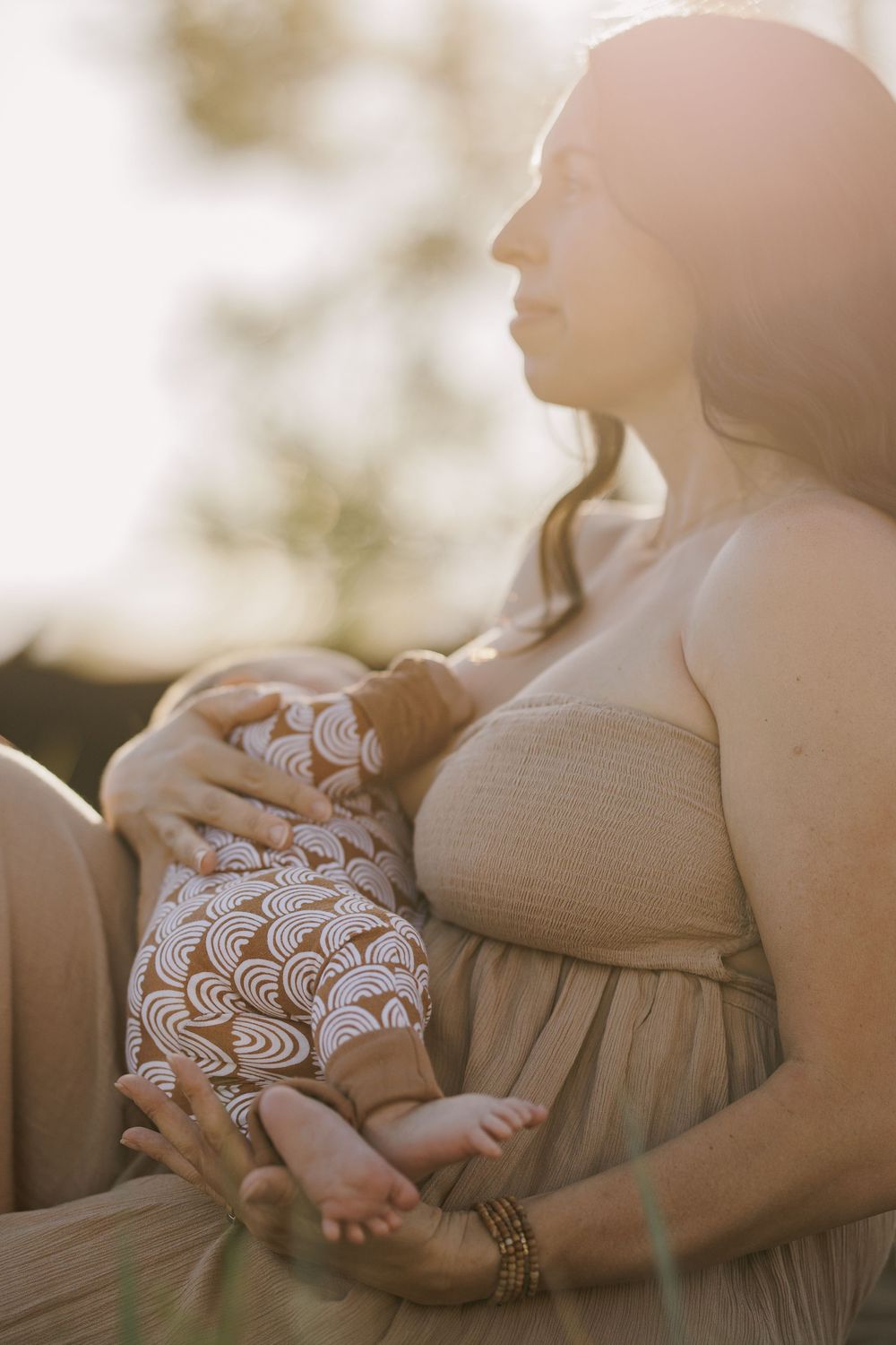 Woman in beige dress holding infant in patterned onesie in warm sunset light.
