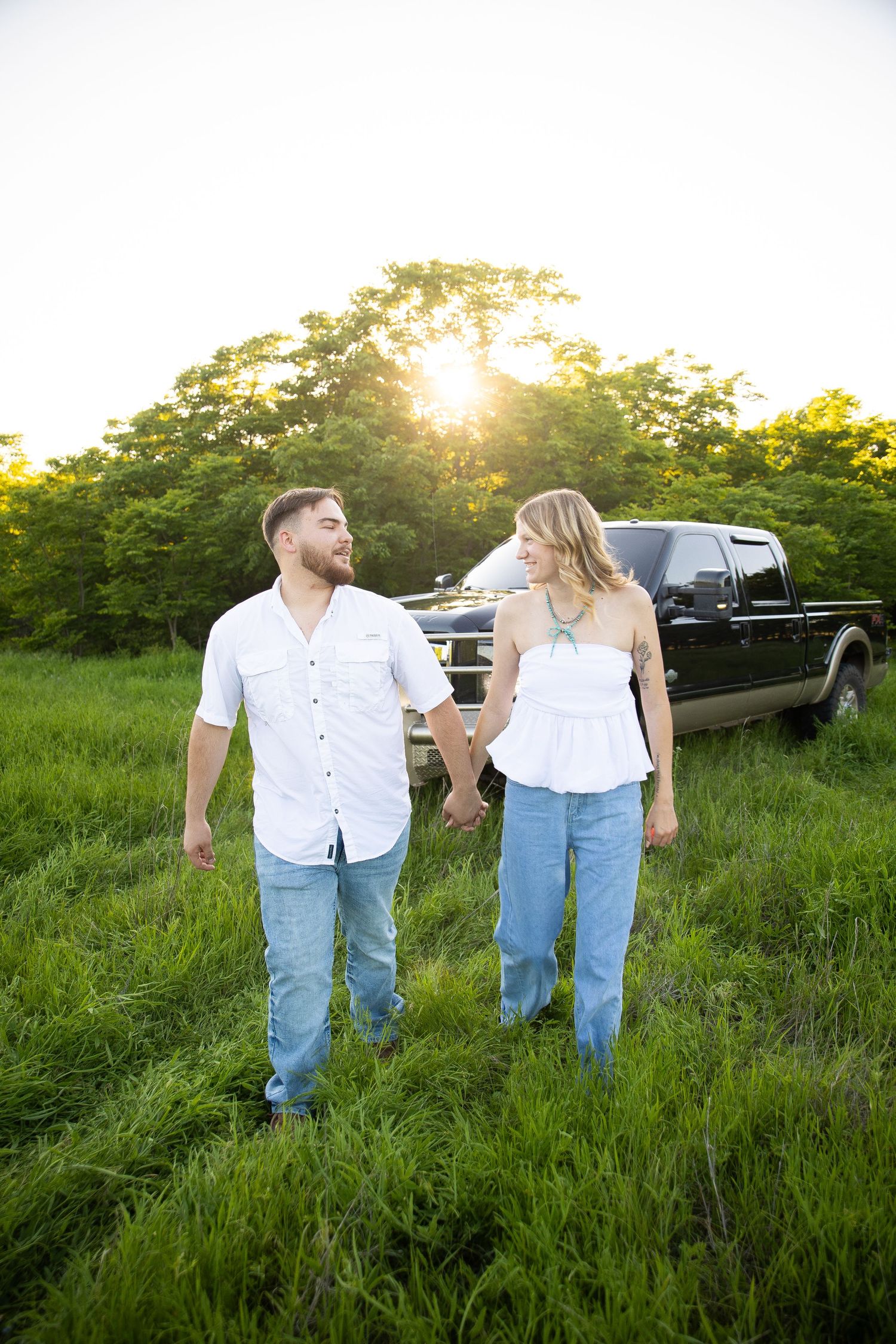 A couple walks hand in hand through a lush green field at sunset while wearing casual white shirts and blue jeans.
