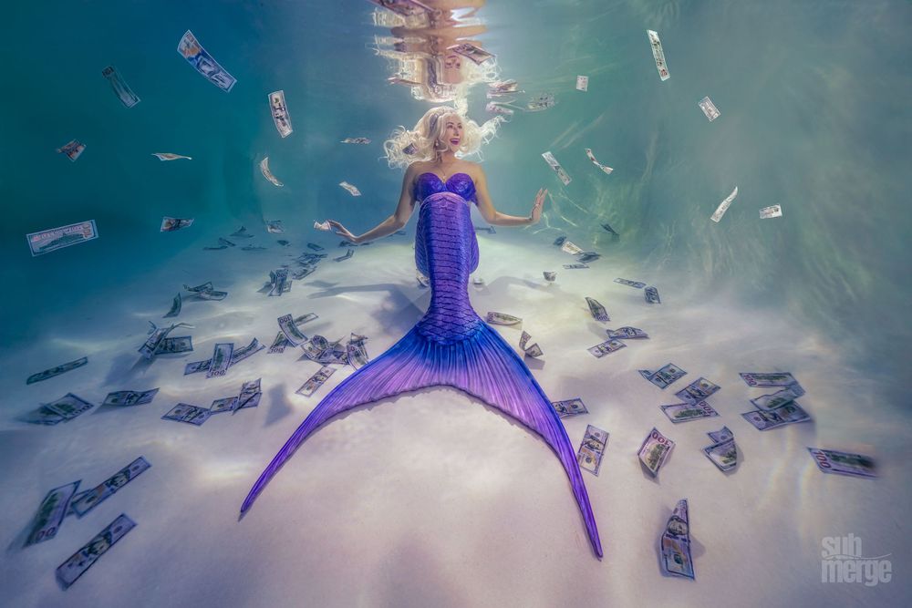 Money floats through turquoise water around a person in a purple dress creating a dreamy underwater scene.