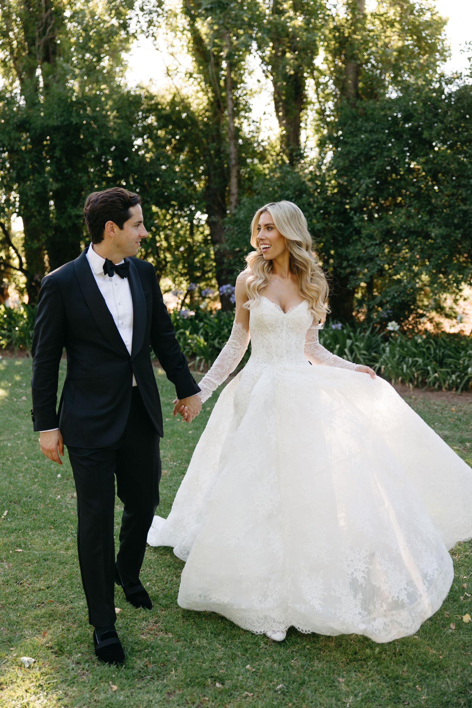 A couple walks hand in hand outdoors, with the bride wearing a flowing white gown and groom in classic tuxedo.