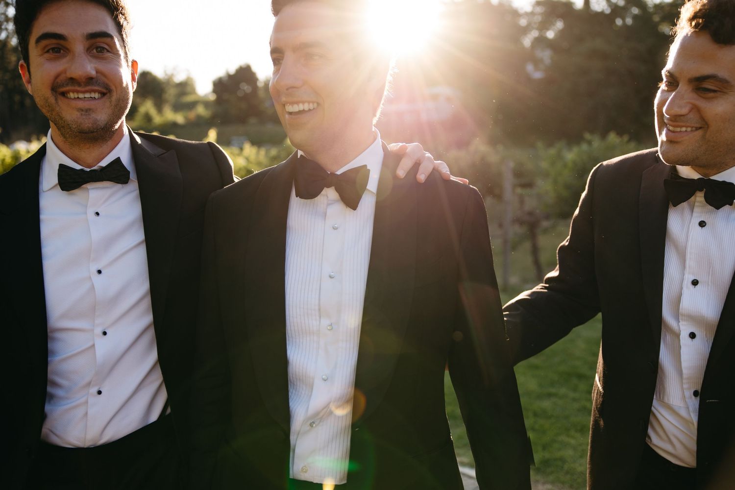 A group in formal black tuxedos and white shirts stand together outdoors during golden hour sunset.