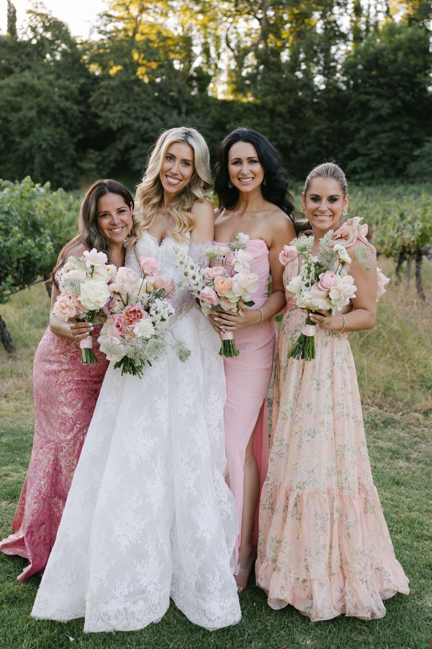 A bridal party in pink and white dresses poses together outdoors holding beautiful floral bouquets.