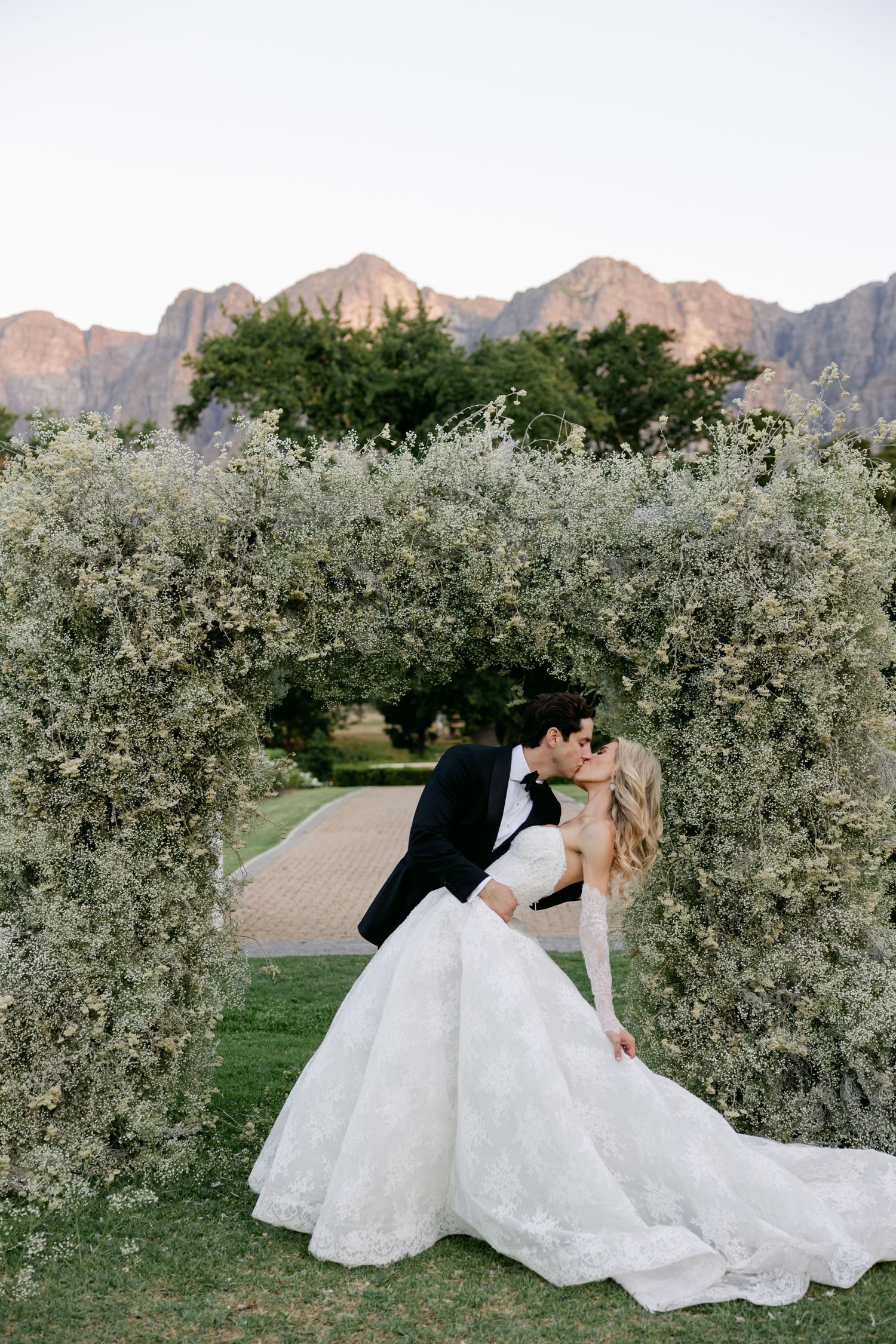 A romantic wedding couple shares an intimate kiss in a blooming orchard with mountain peaks in the background.