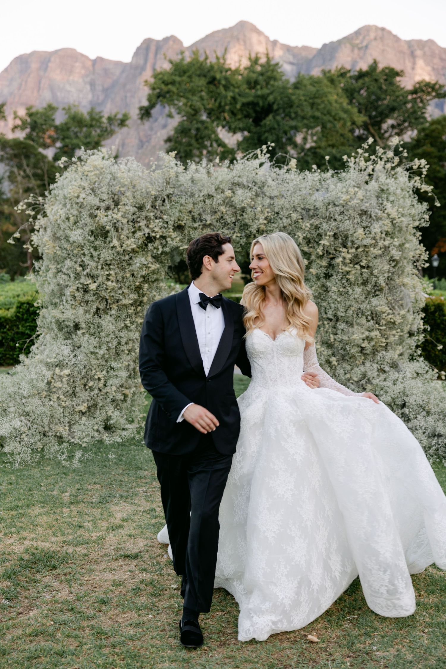 A couple in wedding attire shares a romantic moment in front of a floral arch with mountains in the background.