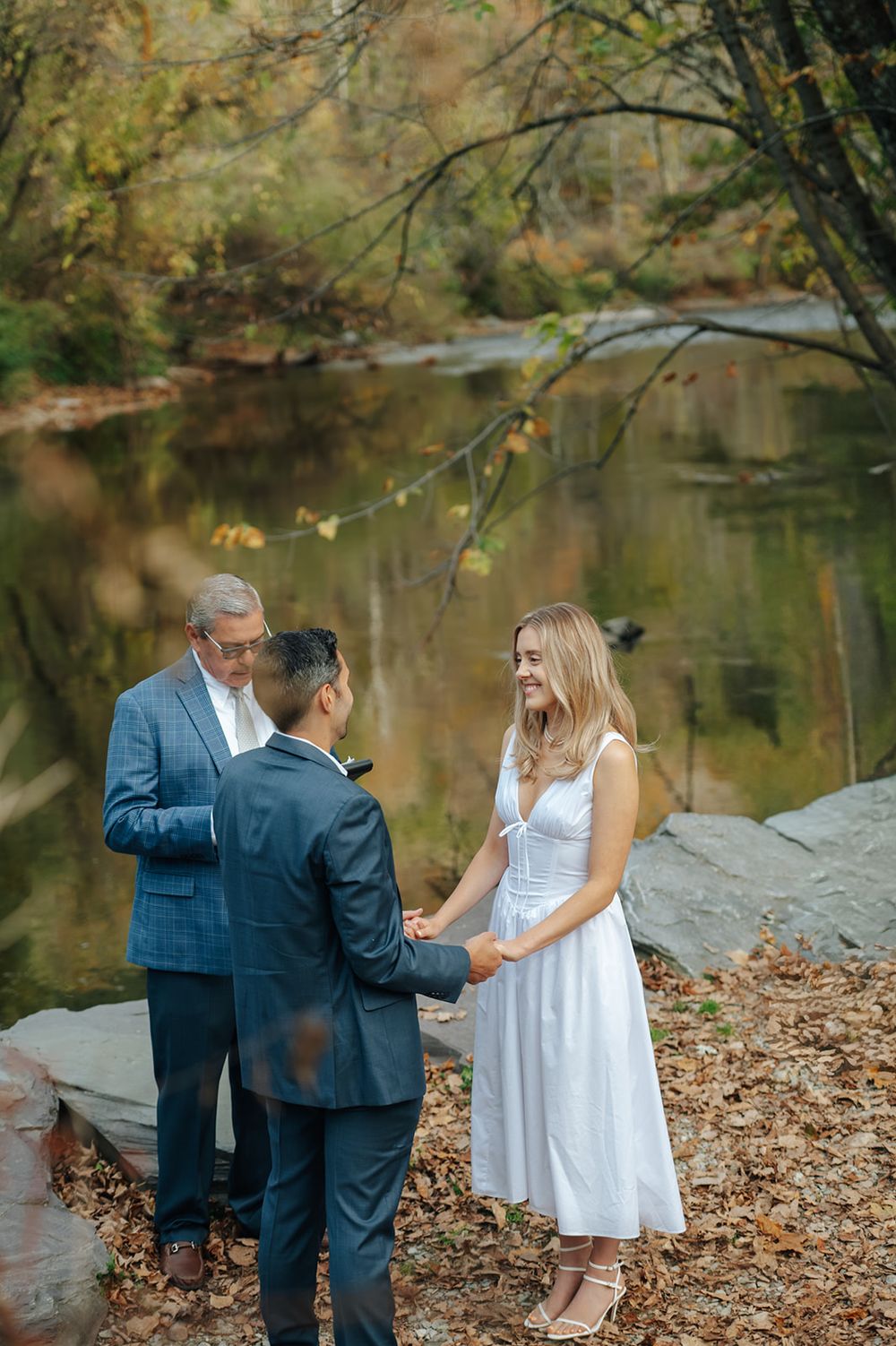 A couple shares a tender moment by a tranquil pond surrounded by autumn foliage and fallen leaves.