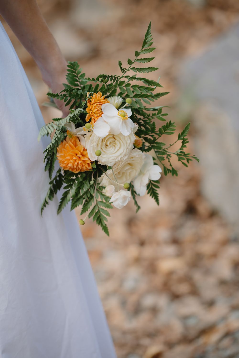 A delicate wedding bouquet detail featuring white roses and orange marigolds with green fern leaves against a light background.