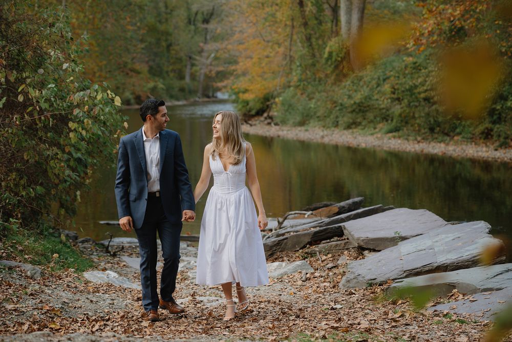A romantic couple strolls along a wooded path near a tranquil lake during an autumn engagement photoshoot.