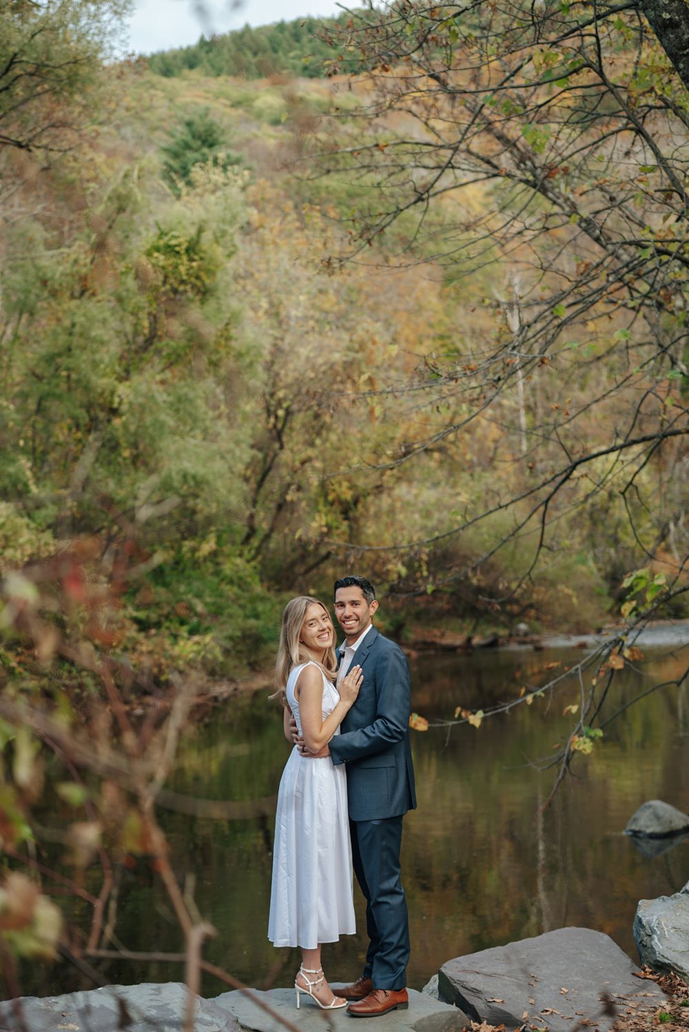 A couple in formal attire stands on rocks by a serene pond surrounded by autumn foliage in a wooded setting.