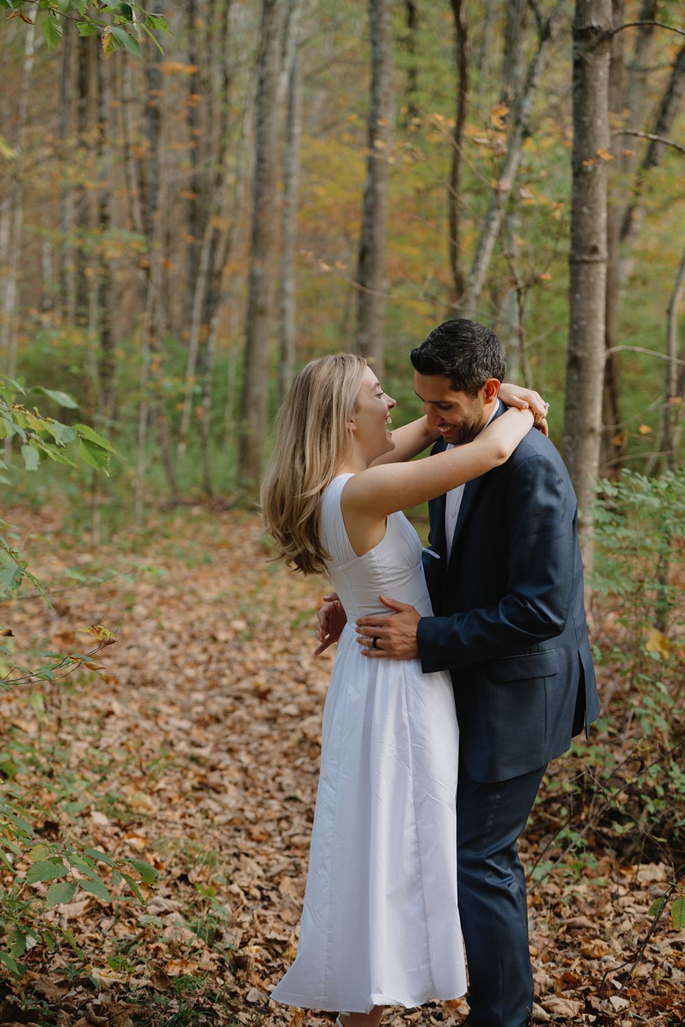 A couple in formal attire dancing and embracing in an autumn forest setting with fallen leaves on the ground.