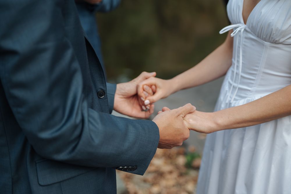 Close-up of couple holding hands during outdoor wedding ceremony, showing white dress and dark suit details.