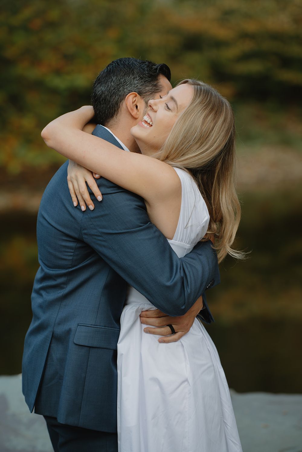 A romantic couple in formal attire sharing an intimate embrace by a tranquil waterfront during an autumn day.